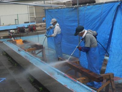 横並びで手袋をしたキャップを被り水色のつなぎ姿の両手に持った高圧洗浄機を使用し木枠の上に置かれた残存部材に脱塩作業を施す地域住民の男性2人の写真