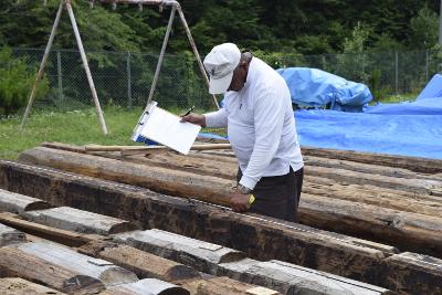 左手に持ったメジャーで残存部材の計測をおこなう右手にバインダーに挟んだ紙とペンを持った男性とその前後に並べられた多数の部材の写真