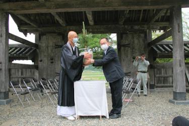 華蔵寺内で宝珠マツのクローン苗木を手渡すマスクにスーツ姿の右側の東北育種場長とそれを受け取るマスクに袈裟姿の左側の住職を後ろから携帯電話で撮影する男性の写真