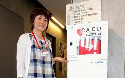 AEDが設置されている区画の前で笑顔を見せる女性職員の写真