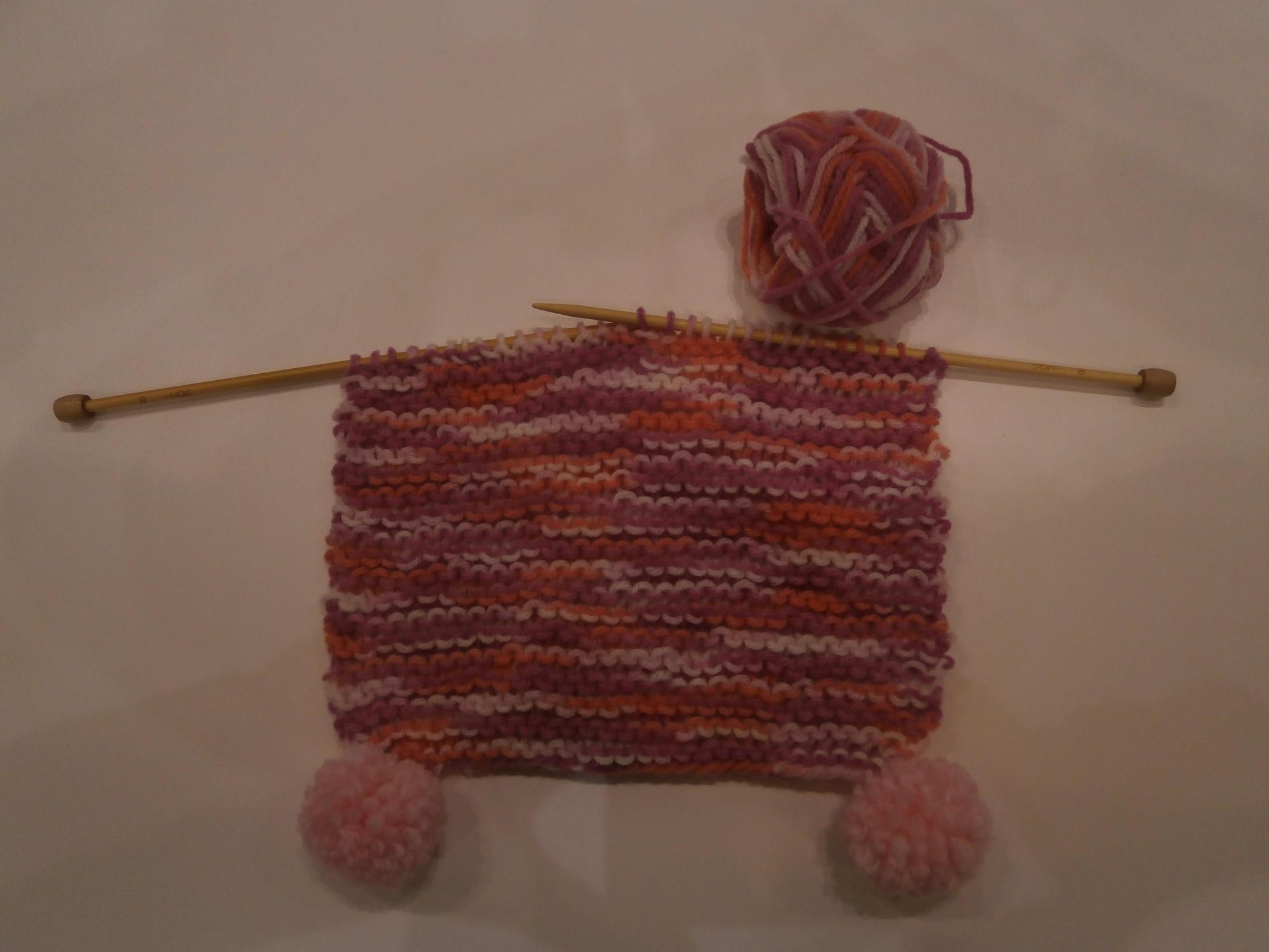 端に黄色のポンポンが二つついた赤と黄色の混じった毛糸で編みかけのマフラーの写真