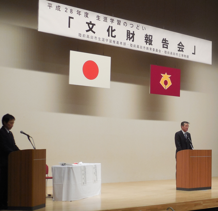 陸前高田市コミュニティホールの一室で文化財報告会と書かれた横断幕の左下に日本国旗が右下に陸前高田市市章が掲げられその両端の壇上に立つスーツ姿の男性の写真