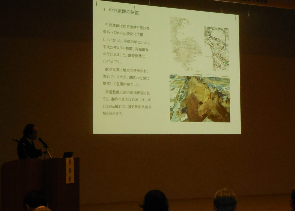 陸前高田市コミュニティホールの一室でプロジェクターを使用しながら壇上で中沢遺跡の発掘記録と出土遺物の整理について説明する市委員会の佐藤氏の写真