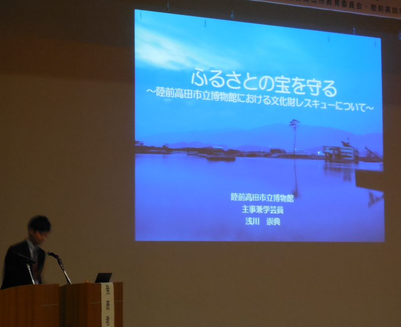 陸前高田市コミュニティホールの一室でプロジェクターを使用しながら壇上で陸前高田市立博物館における文化財レスキューについて説明する市立博物館の浅川氏の写真