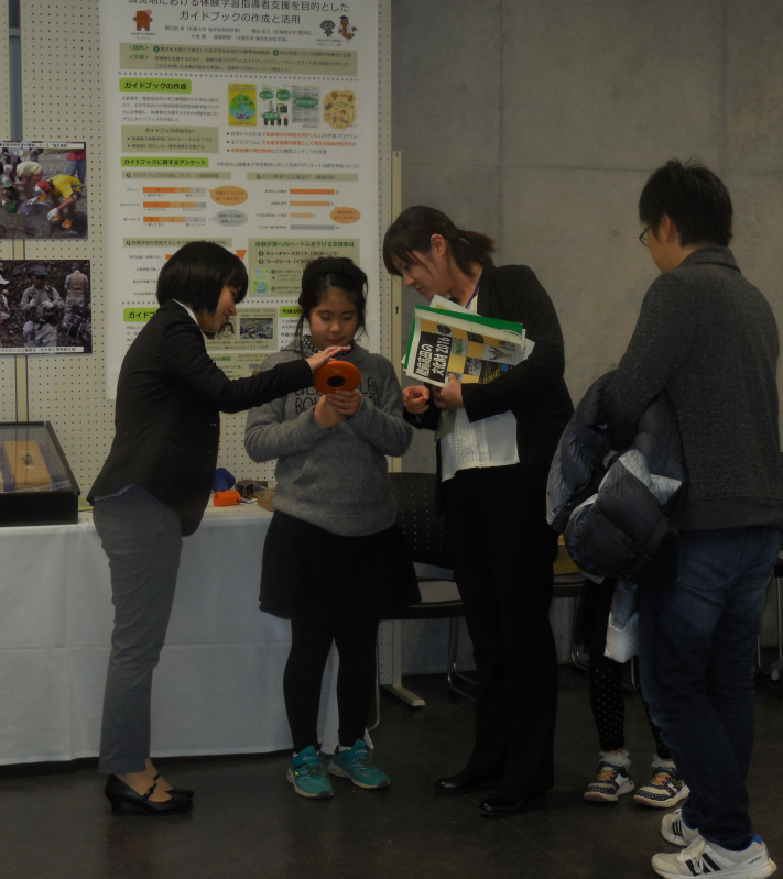 陸前高田市コミュニティホールの一室で女性職員の説明を受けながらオレンジ色の展示物を左手に持つ少女とその様子を左横で覗き込む母親の写真