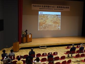 陸前高田市コミュニティホールの一室でプロジェクターを使用しながら壇上で埋蔵文化財調査成果について報告する村木埋蔵文化財センター長の写真