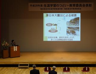 陸前高田市コミュニティホールの一室でプロジェクターを使用しながら壇上で一般文化財事業について説明する教育委員会の福原氏の写真