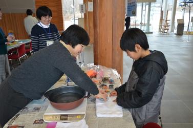 陸前高田市コミュニティホールの一室でおこなわれた体験コーナーで前屈みになりながら少年に説明する女性職員とその様子を微笑みながら見守る女性職員の左横に立つ女性職員の写真