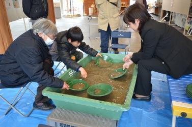 体験コーナーで緑色の水槽に入ったものを緑色の器ですくうパイプ椅子に座る少年と祖母と向かい側のカラフルなベンチに座る女性の写真