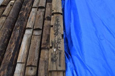 ブルーシートの左横に置かれた黒文字で漢字が書かれた吉田家住宅の残存部材とその左横に並べられた多数の部材の写真