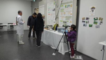 陸前高田市コミュニティホールの一室に展示された数々の文化財を興味深げに眺める男女の来場者らと展示物の望遠鏡をつま先立ちしながら覗き込む少女の写真