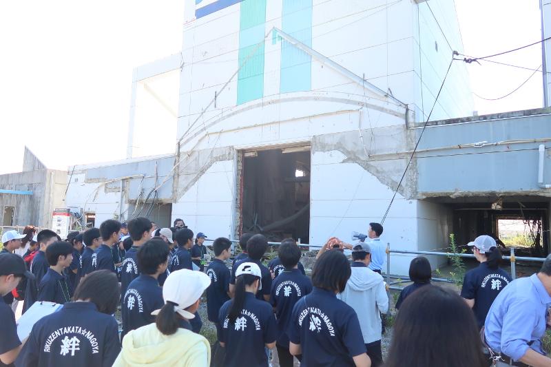陸前高田市にある被災した道の駅高田松原の前で右手にメガホンを持ちながら話す男性の前方で起立して話に耳を傾ける紺色のポロシャツを着た名古屋市の中学生たちの写真
