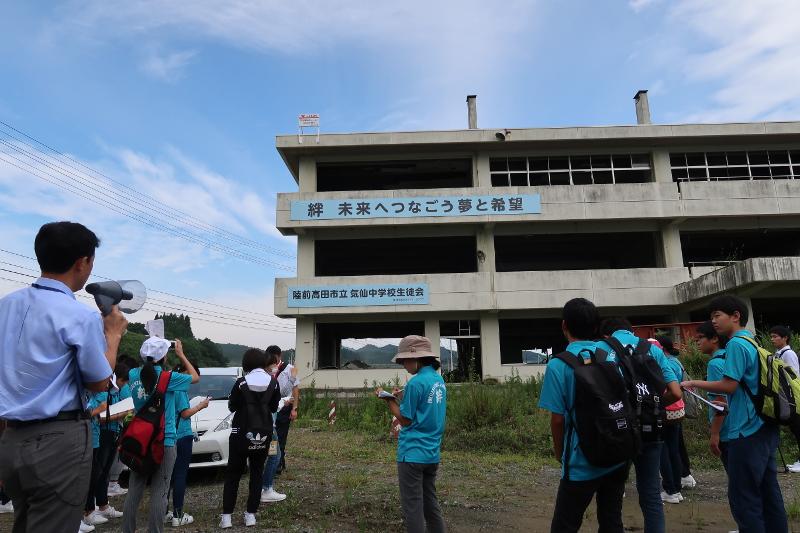 被災した陸前高田市立気仙中学校の前で携帯電話で校舎を撮影したり後方で左手でメガホンを持ちながら話す男性の方を向いている中学生たちと校舎に掲げられた絆未来へつなごう夢と希望と黒字で書かれた水色の横断幕の写真