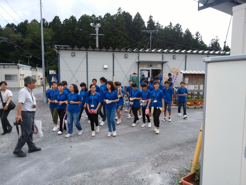 陸前高田市にある仮設住宅をボランティア体験のため訪れる青色のポロシャツを着た名古屋市の中学生たちの前を歩く右手にカメラを左手に紙袋を持った男性の写真