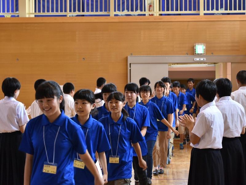 陸前高田市立第一中学校の体育館で両端に並んで拍手で迎える生徒たちの間をはにかみながら歩く青色のポロシャツを着た名古屋市の中学生たちの写真