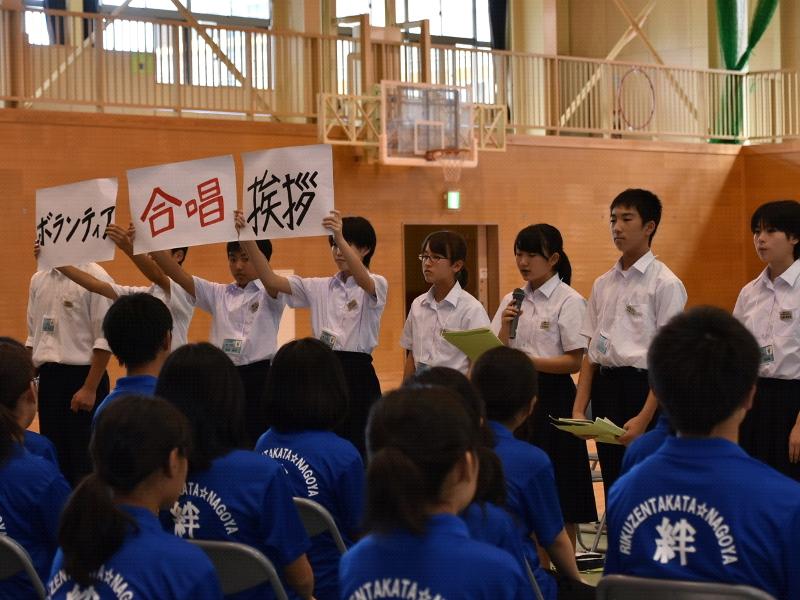 右側から黒字でボランティアと書いた紙と赤字で合唱と書いた紙と黒字で挨拶と書いた紙をそれぞれ両手に持ち掲げる3人の生徒と右手に持ったマイクで喋る女子生徒の前方で話を聞く名古屋市の中学生たちの写真