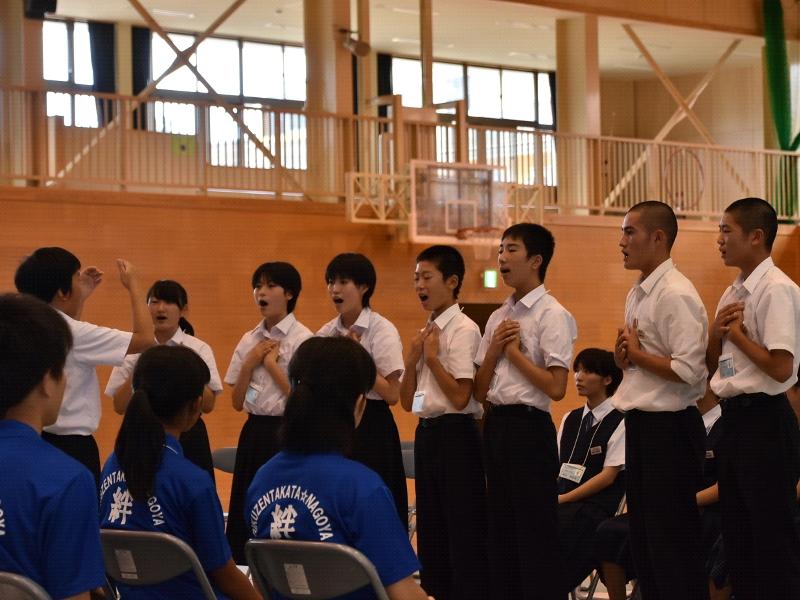 両手を胸の前に置き合唱する生徒たちとその前方で指揮をする男子生徒の後ろでその様子を眺めている青色のポロシャツを着た名古屋市の中学生たちの写真