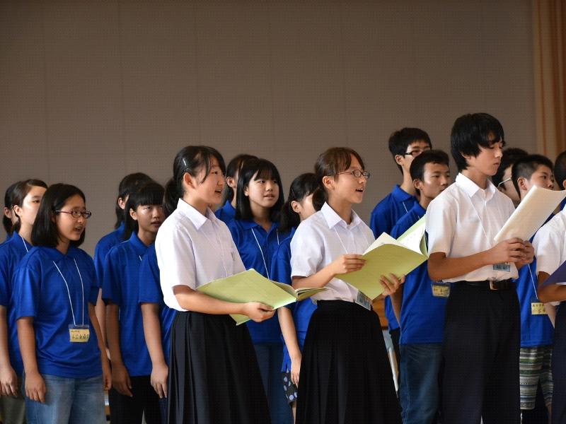陸前高田市立第一中学校の体育館で両手に紙を持ちながら合唱する生徒たちの後ろで起立し横並びで同じように合唱する青色のポロシャツを着た名古屋市の中学生たちの写真