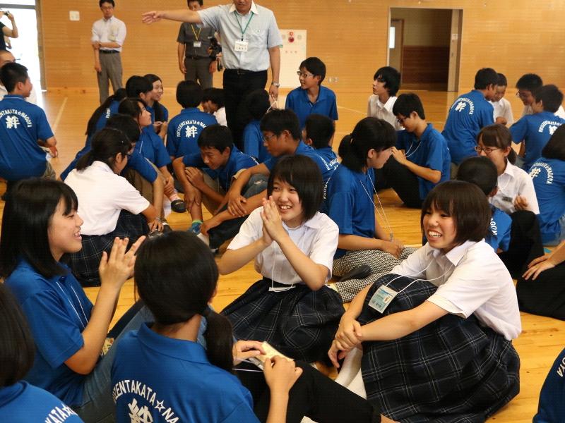 陸前高田市立第一中学校の体育館の床に数人一グループになって座りお互いに交流を深める生徒と青いポロシャツを着た名古屋市の中学生たちの写真