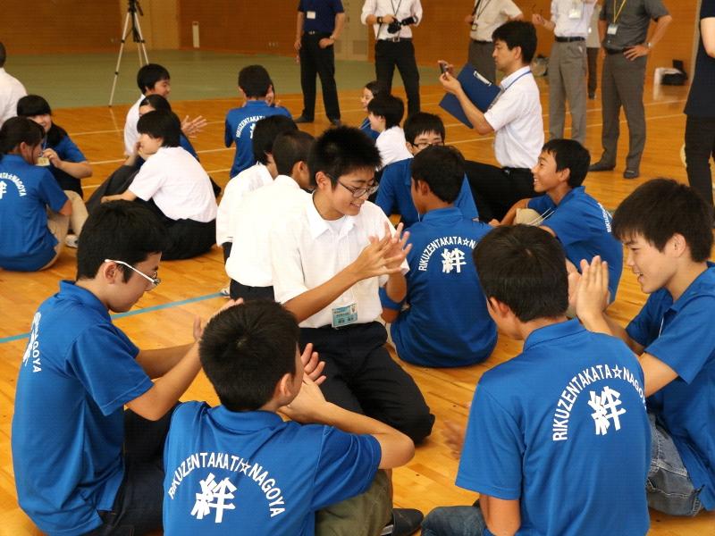陸前高田市立第一中学校の体育館の床に数人一グループになって座りお互いに交流を深める生徒と名古屋市の中学生たちの様子を見守る関係者らの写真