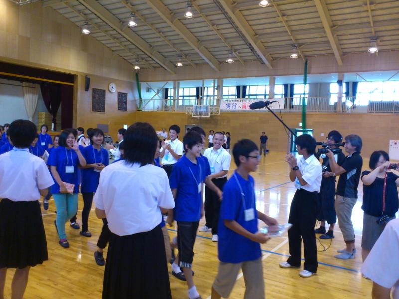 両端に並んで拍手で見送る生徒たちの間を笑顔で歩く青色のポロシャツを着た名古屋市の中学生たちの様子をプロ用のカメラで撮影する男性とその右横でヘッドフォンをして音声マイクを持つ男性写真