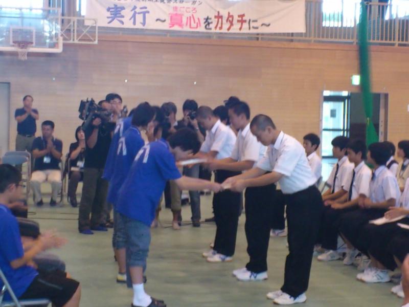 左側の生徒たちと右側の名古屋市の中学生たちが頭を下げながら横並びで物を手渡している様子を同じように横並びでパイプ椅子に座りながら拍手している右端の生徒たちと左端の中学生たちの写真