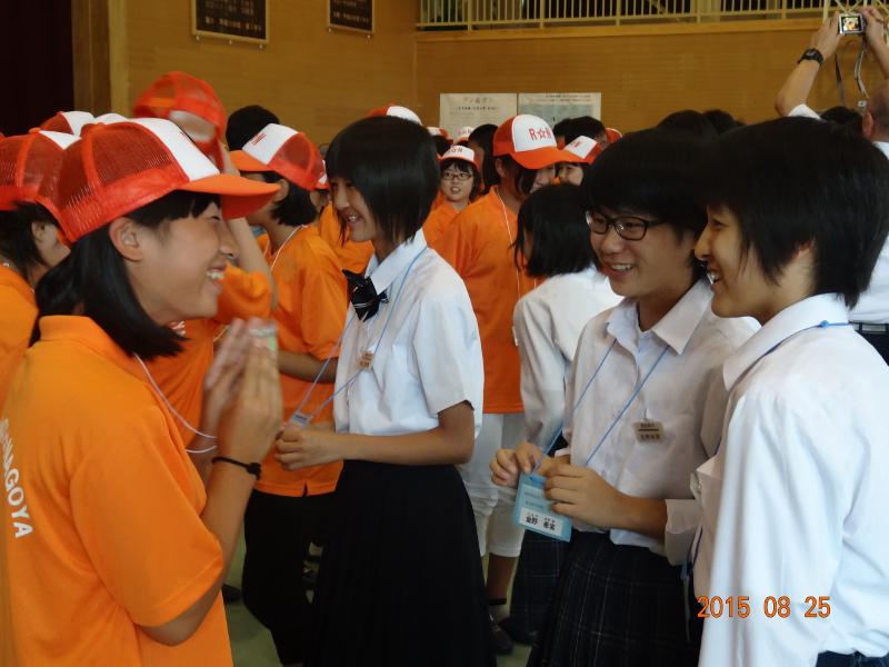 陸前高田市内の中学校の体育館で首から下げた名札をお互い笑顔で見せ合う陸前高田市の中学生たちとオレンジ色のキャップにオレンジ色のポロシャツを着た名古屋市の中学生たちの写真