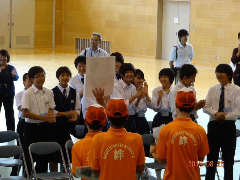 陸前高田市の中学校の体育館で右手に紙を持ちそれを掲げて見せる名古屋市の男子中学生と後ろで左手にマイクを持つ男子中学生の向かい側で起立して笑顔を浮かべながら拍手する陸前高田市の中学生たちの写真