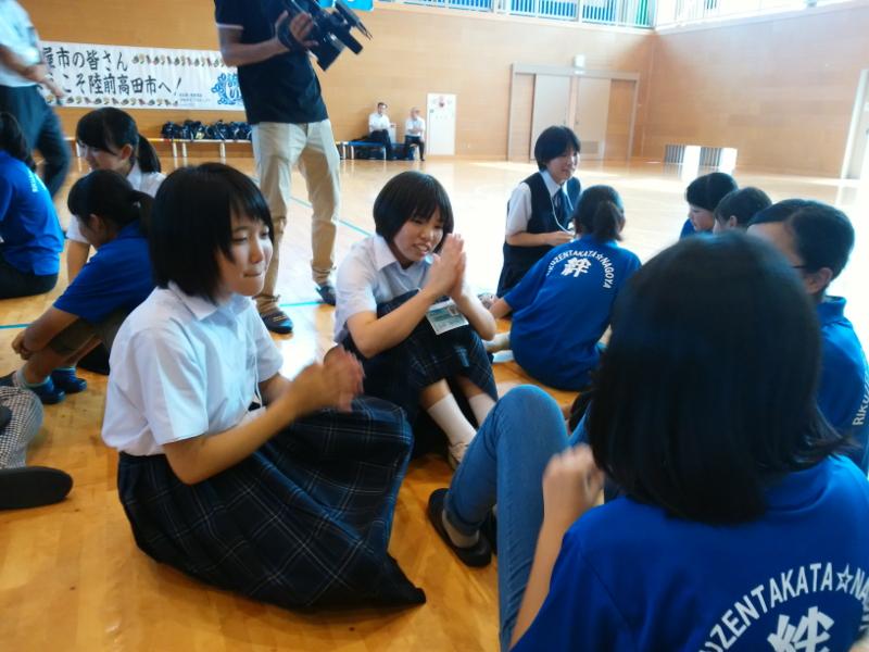 陸前高田市立第一中学校の体育館の床に数人一グループになって座りお互いに交流を深める女子生徒と名古屋市の女子中学生たちの様子をプロ用のカメラで上から撮影する男性の写真
