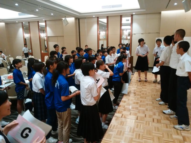 壇上に横1列で並び楽しそうな表情を浮かべる陸前高田市の中学生たちとその下で壇上の方に群がって笑顔で起立しながら話を聞いている中学生たちの写真