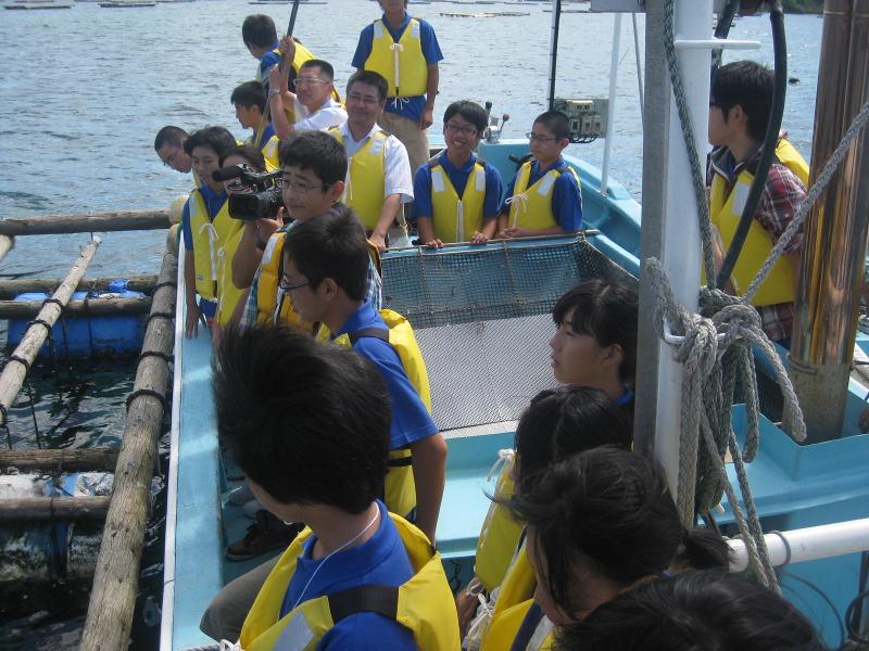 いかだ式養殖ワカメの右横で船に乗りながら海の中のワカメを覗く青色のポロシャツの上に黄色のライフジャケットを着た中学生らの写真