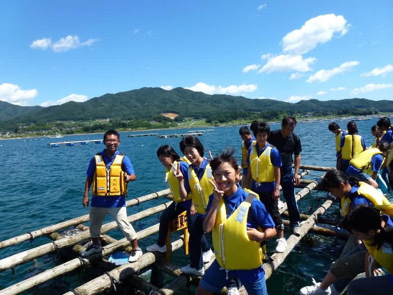 青空の下広大な山と海をバックに木製のいかだの上に乗りカメラに向かってピースをしたり海の中のワカメを覗き込む青色のポロシャツの上に黄色のライフジャケットを着た中学生らの写真