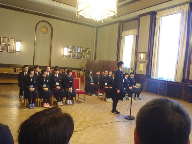名古屋市役所の正庁で起立しスタンドマイクで挨拶をする陸前高田市の代表者の女子中学生とその後ろで縦2列横並びに座る中学生たちの写真