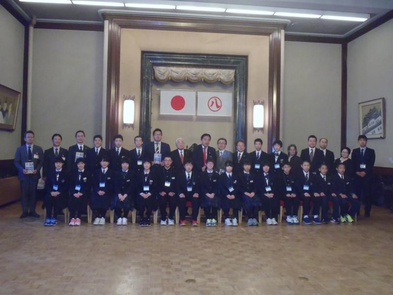 名古屋市役所の正庁で右側に掲げられた日本国旗と左側に掲げられた名古屋市の市章の下で前列の中学生たちは座り後列の中学生らは起立して記念撮影をする写真