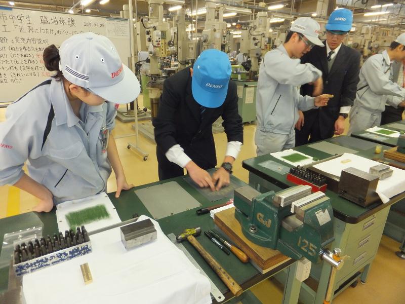 白文字でDENSOと刺繍された水色のキャップを被った陸前高田市の男子中学生2人とその右横で職場体験の指導をする赤文字でDENSOと刺繍された灰色のキャップを被って制服を着たの社員3人の写真