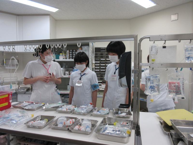 研修中と書かれた水色の腕章をしたマスクに看護師服姿の陸前高田市の女子中学生2人が白色の台に置かれバットに入った医療用具を下向きで見ている左横でマスク姿で丁寧に説明する女性看護師の写真