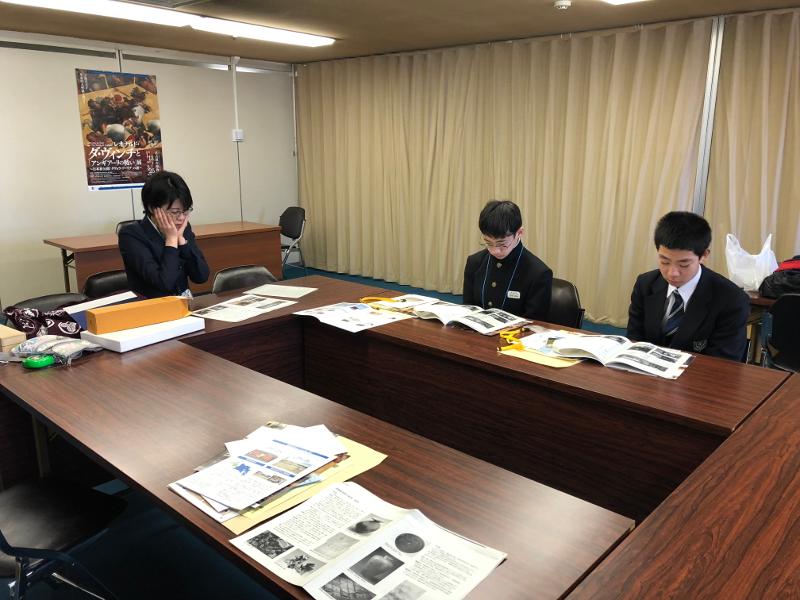 茶色の会議用横長机が角ばったO型に4台置かれその正面側で机に置いた資料を両手で両頬を包む陸前高田市の女子中学生と左側で資料を読む2人の男子中学生の写真