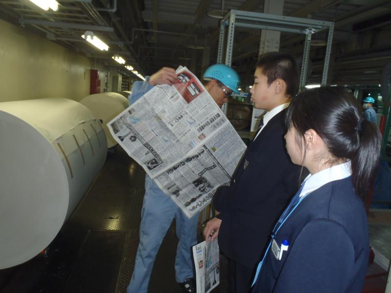 水色のヘルメットを被り新聞紙を広げ説明をする男性とその左横で両手を前に組み左手で新聞紙を持つ男子中学生とその右横の女子中学生が前方にある巨大ロール紙を見ている写真