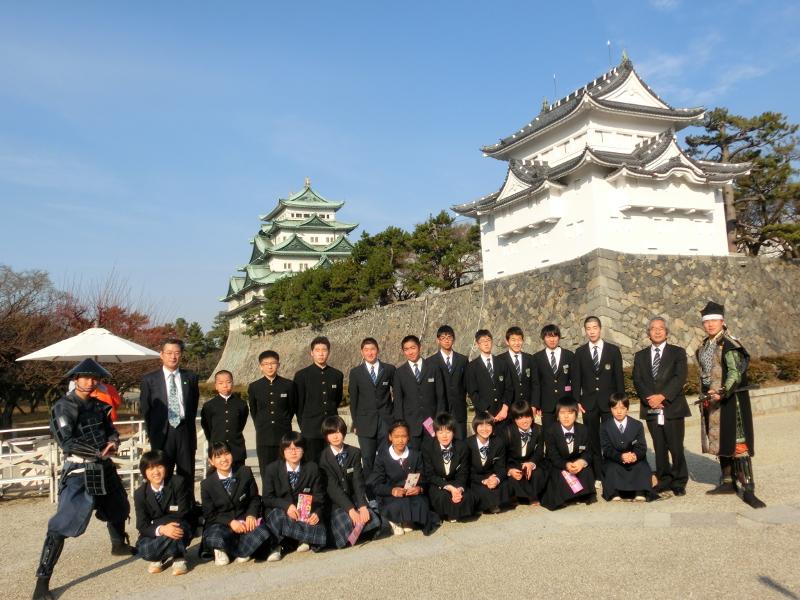 青空の下名古屋城をバックに前列の女子中学生はしゃがんで後列の男子中学生らは起立して横に並びその左端に立つ武将姿の男性と右端に立つ足軽姿の男性と記念撮影をする写真