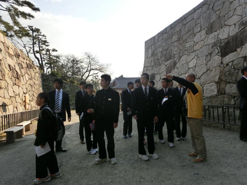 名古屋城の石垣の前で左手の人差し指を使い説明をする黄色い法被を着た観光案内ボランティアの男性と男性が指さした方向を向きながら話に耳を傾ける男子中学生らの写真