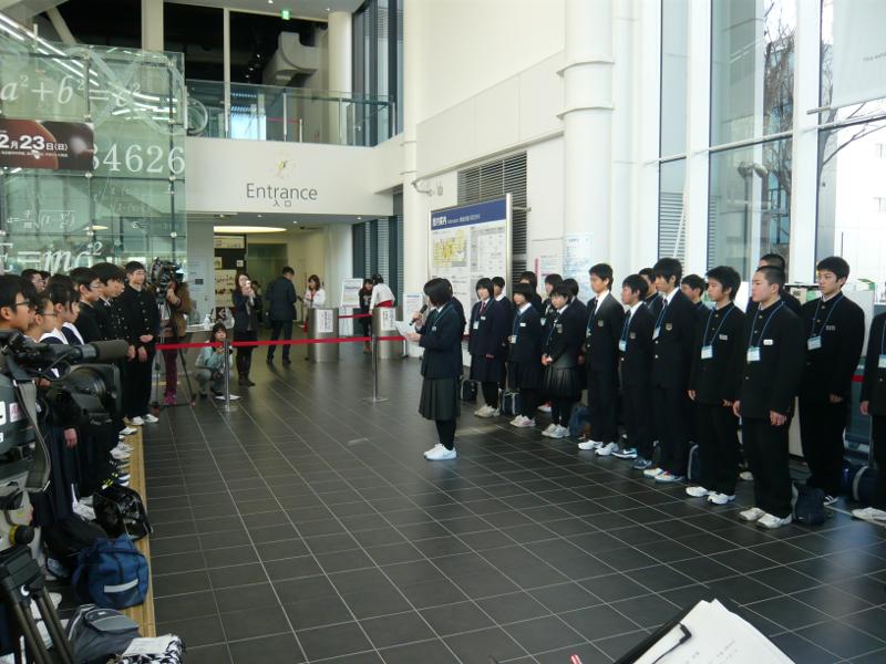 建物の入り口で前方に横並びに並んでいる名古屋市の中学生たちに対して右手にマイクを持ち左手に紙を持ちながら挨拶をする陸前高田市の女子中学生とその後ろで縦2列に横に並ぶ中学生たちの写真