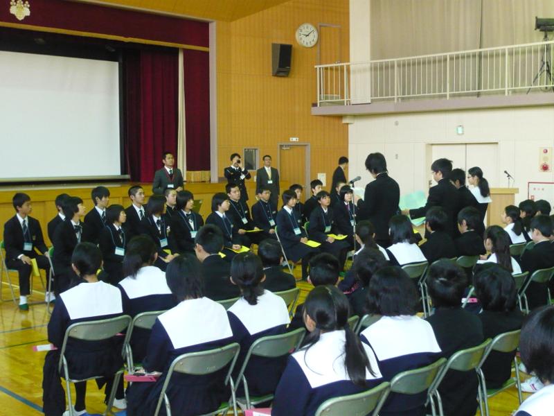 名古屋市立山王中学校の体育館で前方に縦2列で横並びにパイプ椅子に座る陸前高田市の中学生たちに対して歓迎の挨拶をする男子生徒とその後ろで横並びにパイプに椅子に座る生徒たちの写真