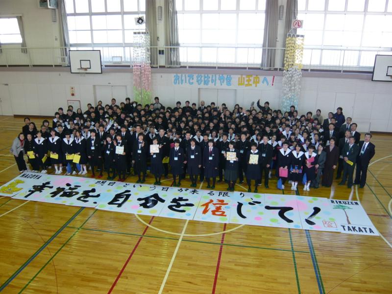 体育館で右側に陸前高田市の市章が左側に名古屋市の市章がてっぺんに飾られた二つの大きな紙飾りの間においでなはりやせ山王中へと書かれた横断幕の下で記念撮影をする中学生らの前に置かれた幸福を自分を信じてと書かれた紙を上から撮影した写真