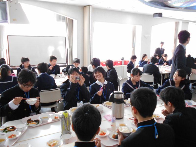 数人一グループになり出された給食を食べながら会話をする中学生たちとその様子を見守る男性教師の写真