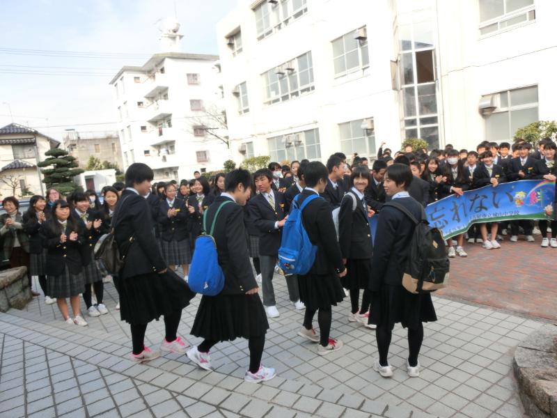 名古屋市立城山中学校内で青色の横断幕を持ちながら拍手で見送る生徒らと恥ずかしそうな表情を浮かべながら見送られる陸前高田市のリュックを背負った女子中学生5人の写真