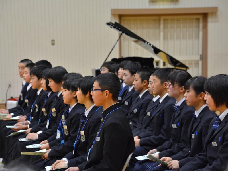 名古屋市立丸の内中学校の体育館で縦2列で横並びにパイプ椅子に座り真剣な表情で前方を向いている陸前高田市の中学生たちの奥にあるグランドピアノの写真