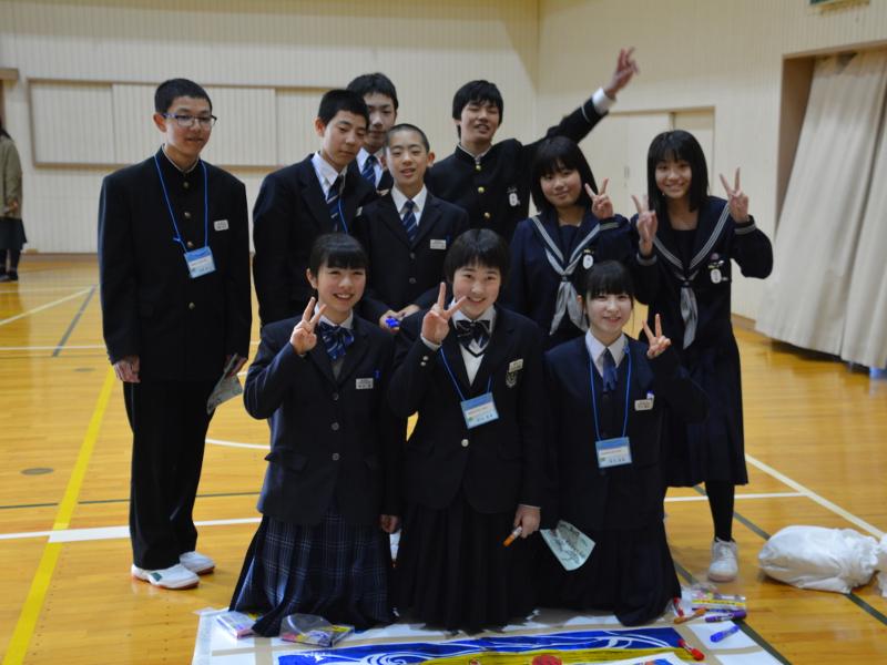 名古屋市立丸の内中学校の体育館で横並びに1列目の3人の女子中学生は膝立ちで2列目と3列目の中学生たちは起立して記念撮影をする中学生たちの写真