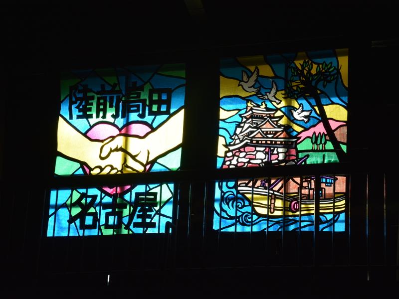 陸前高田名古屋と書かれた間にピンク色のハートマークを背景に互いに握手を交わす手が描かれた左側のステンドグラスと右側の名古屋城と屋形船が描かれたステンドグラスの写真