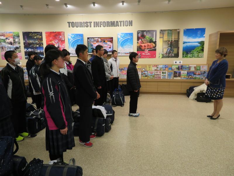 観光案内所内でTOURISTINFORMATIONの文字看板の下で横並びに整列する陸前高田市の中学生たちと1人だけ一歩前に出ている代表者の男子中学生とその前方で両手を前に置き起立する女性の写真