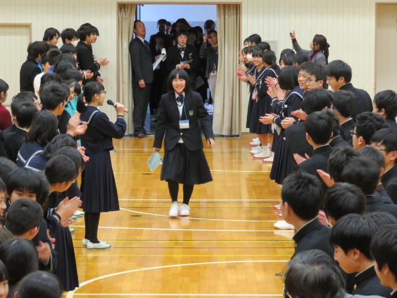 中学校の体育館で両端に集まって拍手で迎える生徒たちと恥ずかしそうに真ん中を歩く陸前高田市の女子中学生とその斜め右前方で右手でマイクを持っている進行役の女子生徒の写真